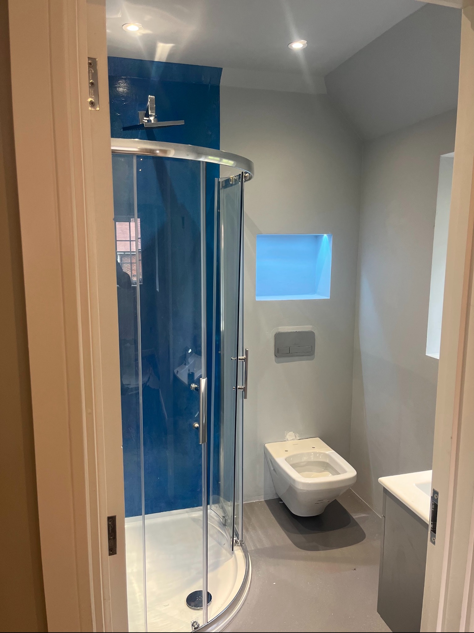 Nowoczesna łazienka z kabiną prysznicową, toaletą podwieszaną i szarą posadzką. Niebieski akcent na ścianie pod prysznicem i wnęka z oświetleniem.