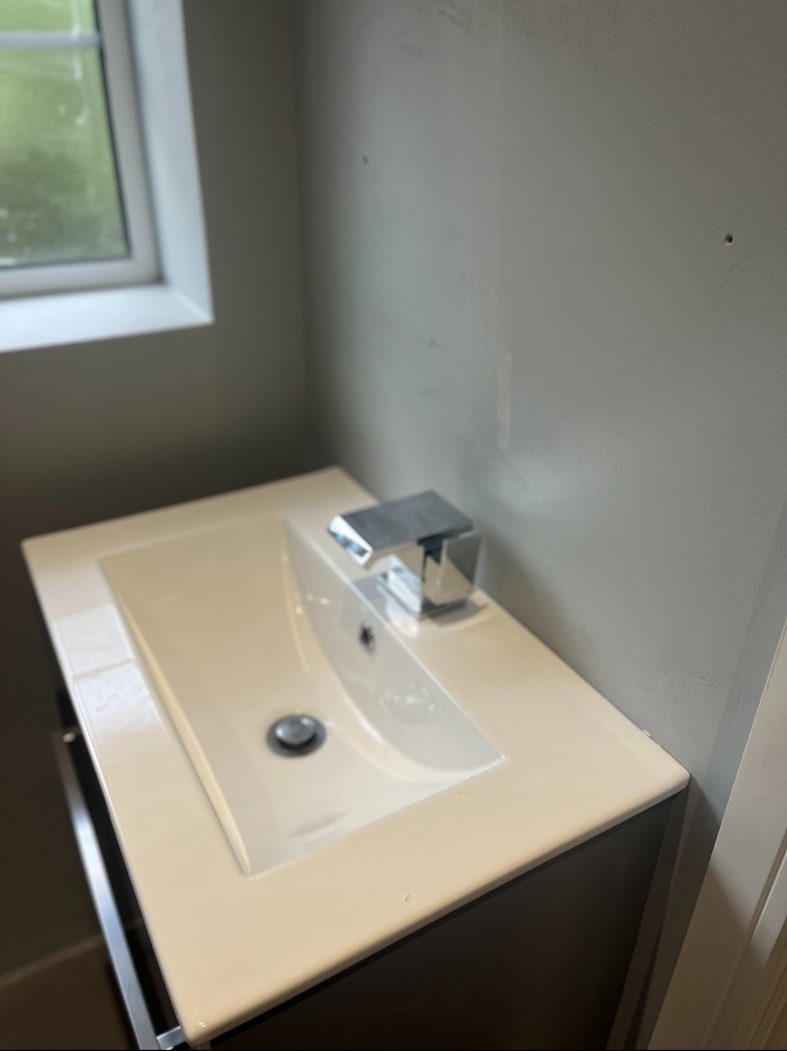 Nowoczesna, biała umywalka z chromowaną baterią, zamontowana na ciemnej szafce w łazience z szarą ścianą i oknem. Minimalistyczny design.