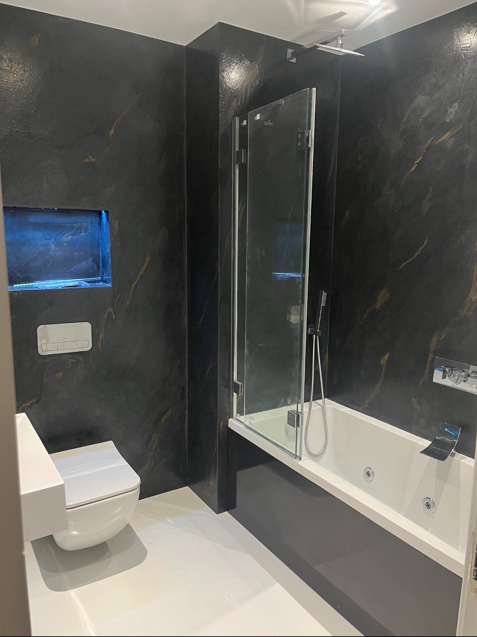 Elegancka łazienka z czarnymi ścianami o teksturze przypominającej marmur, białą armaturą i podświetlaną wnęką. Nowoczesny design z kabiną prysznicową nad wanną.