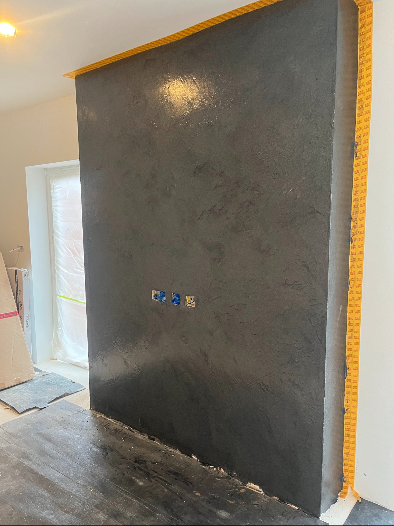 Ściana pokryta szarą, błyszczącą powłoką dekoracyjną, zabezpieczona taśmą malarską. Widoczne puszki elektryczne i fragment podłogi z folią ochronną.