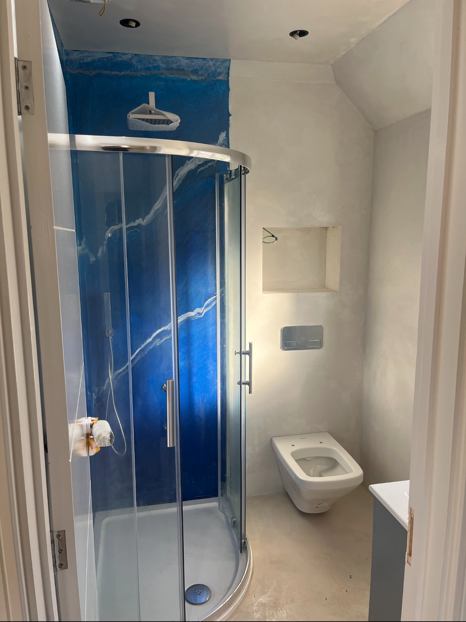 Wykończona łazienka z kabiną prysznicową z niebieską ścianą imitującą marmur i toaletą podwieszaną. Ściany wykończone w jasnym odcieniu szarości.