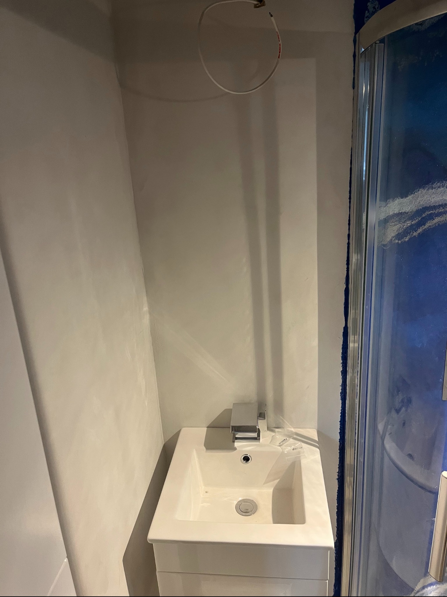 Wykończona łazienka z umywalką i kabiną prysznicową. Ściany pokryte jasnym, gładkim wykończeniem, przypominającym mikrocement. Widoczne detale armatury i oświetlenia.