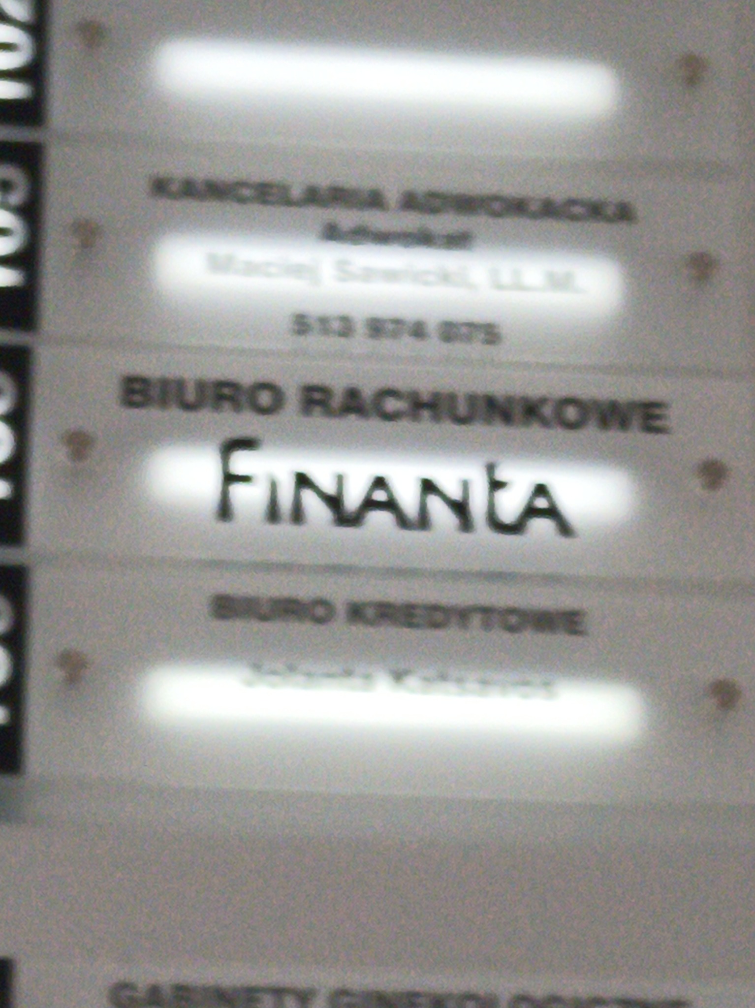 Tabliczka informacyjna na ścianie budynku z napisem 'Biuro Rachunkowe Finanta', obok inne biura: Kancelaria Adwokacka i Biuro Kredytowe. Nierównomierne oświetlenie.
