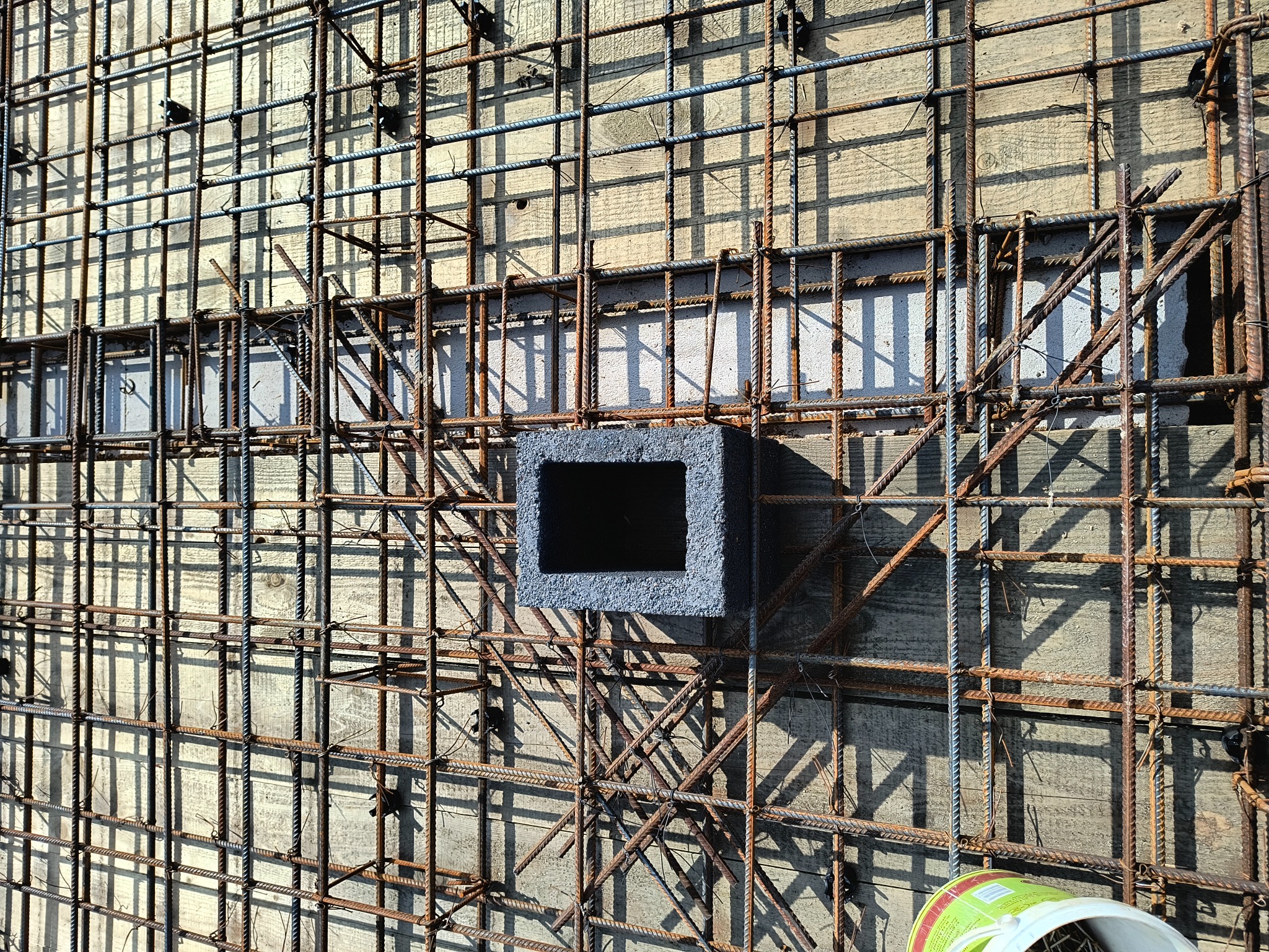 Zbrojenie ściany z otworem na instalacje, widoczne drewniane szalunki i wiązania drutem. Elementy zbrojenia częściowo pokryte rdzą, oświetlone słońcem.