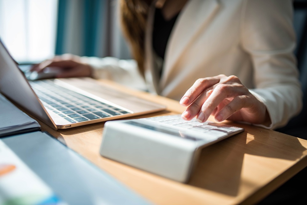 Kobieta w jasnym żakiecie pracuje przy biurku z laptopem i kalkulatorem, wykonując obliczenia. Jasne, naturalne światło pada na biurko.