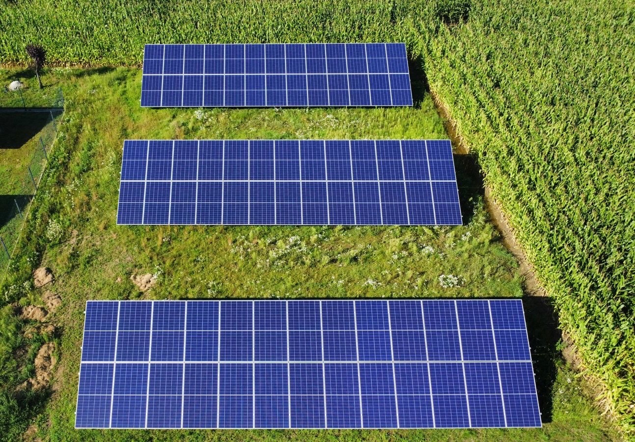 Widok z góry na trzy rzędy paneli słonecznych na zielonej trawie, obok pola kukurydzy. Panele ustawione równolegle, tworząc geometryczny wzór.