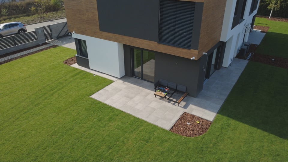 Nowoczesny dom z tarasem wyłożonym jasnymi płytami, widok z góry. Na tarasie meble ogrodowe, obok trawnik i rabata z korą. Minimalistyczny design.