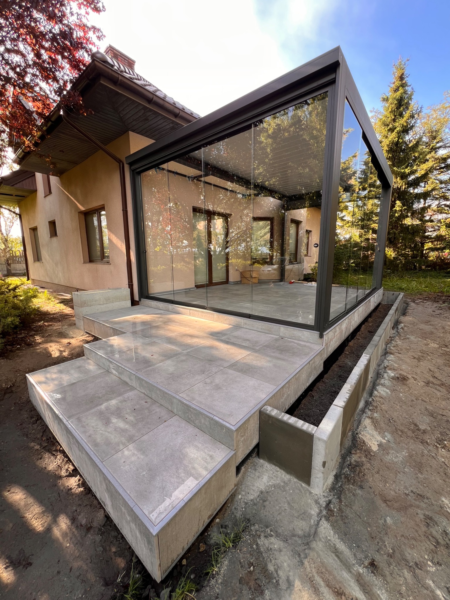 Szklana zabudowa tarasu z szarymi płytkami i schodami, widok z zewnątrz. Nowoczesna konstrukcja z ciemnymi ramami, przylegająca do budynku. Widoczne elementy ogrodu.