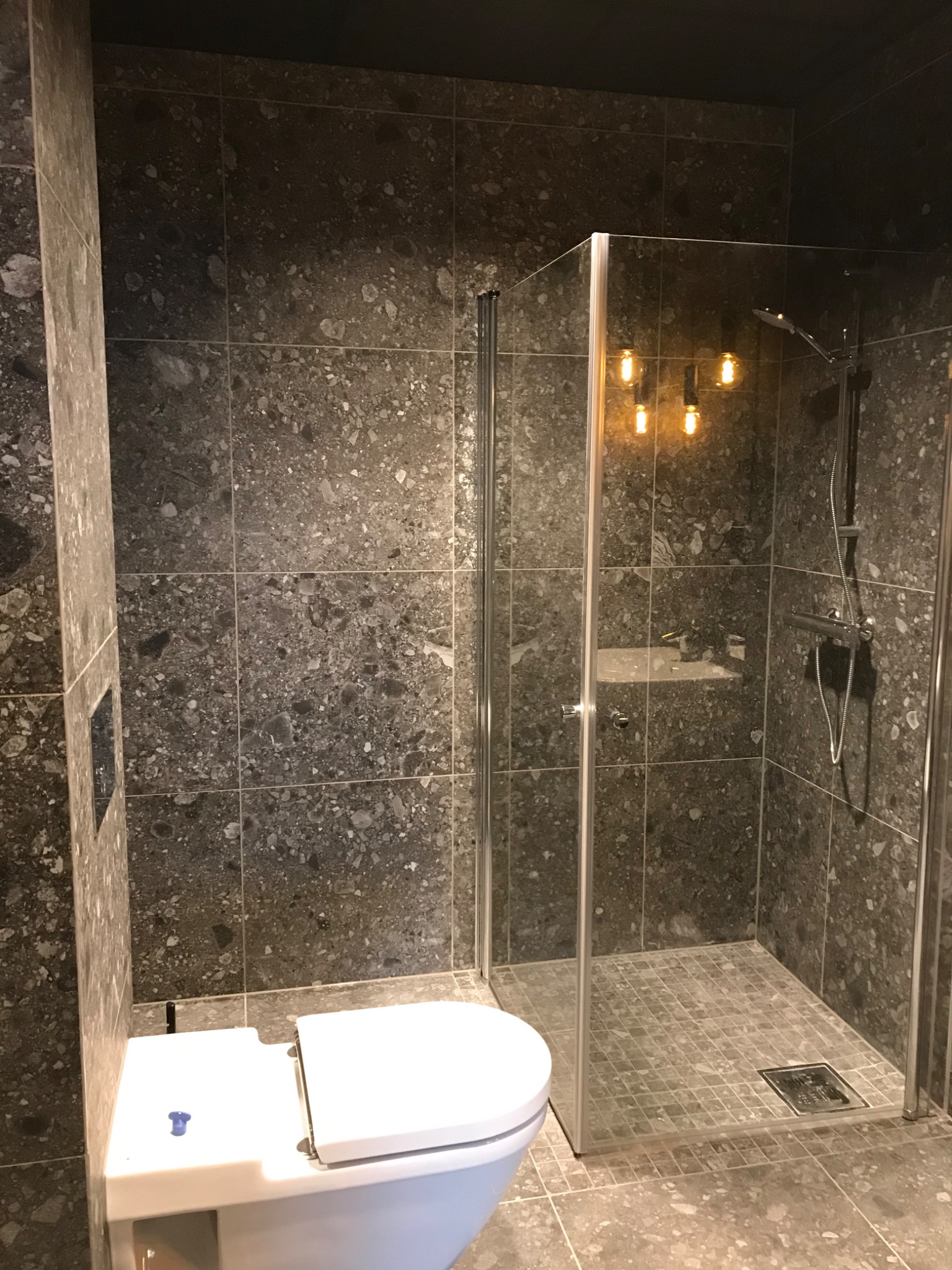 Nowoczesna łazienka z kabiną prysznicową typu walk-in, wykończona szarymi płytkami imitującymi kamień. Widoczny biały sedes i oświetlenie w stylu industrialnym.