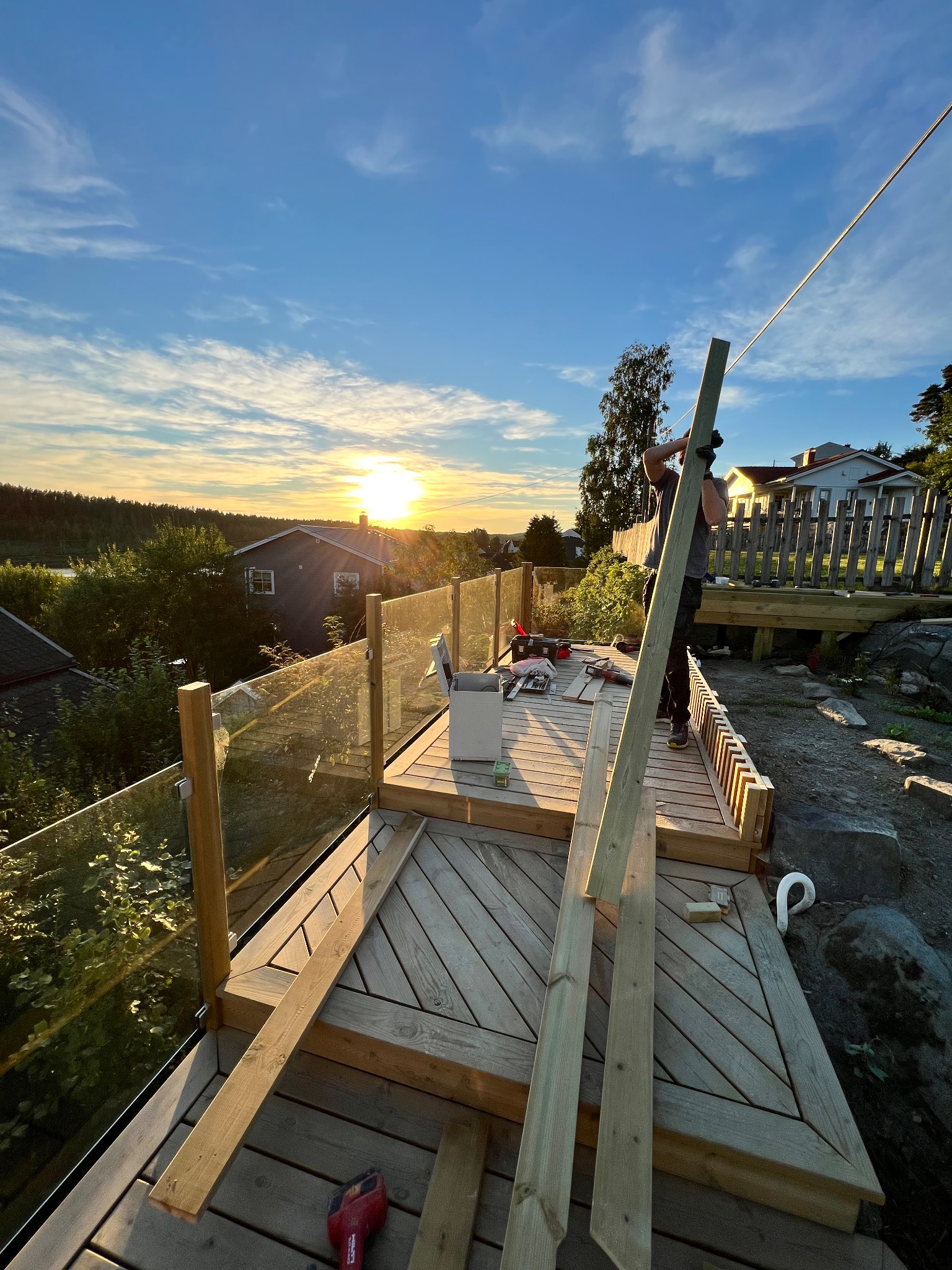 Budowa tarasu drewnianego z przeszkloną balustradą w promieniach zachodzącego słońca. Widoczne narzędzia i deski, częściowo ukończona konstrukcja.