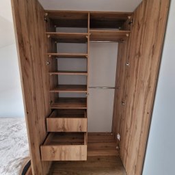 Tomasz Piechota - Otwarta szafa wnękowa z drewnianymi półkami i szufladami, widoczna z perspektywy podłogi, ukazująca wnętrze i strukturę drewna. Widoczne zawiasy i elementy montażowe.