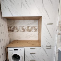Tomasz Piechota - Biała zabudowa meblowa z marmurowym wzorem, integrująca pralkę. Blat drewniany, płytki z motywem liści palmowych. Nowoczesne uchwyty, minimalistyczny design.