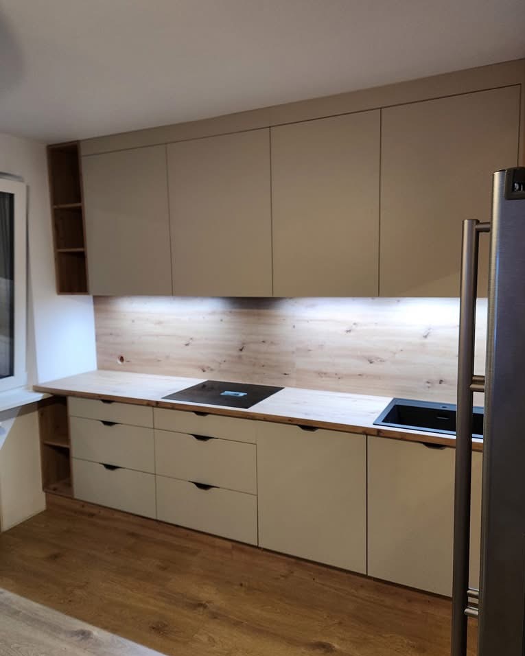 Jasna kuchnia z drewnianym blatem i frontami w odcieniach beżu. Górne szafki sięgają sufitu, a podświetlenie LED dodaje ciepła. Minimalistyczne uchwyty i nowoczesna płyta indukcyjna.