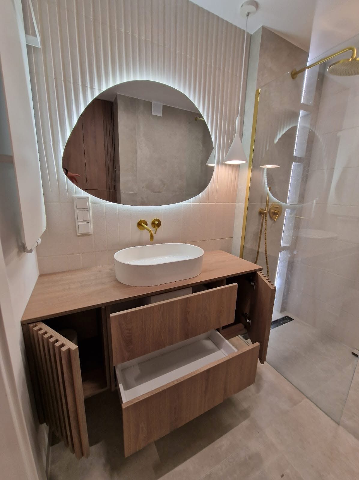 Nowoczesna łazienka z drewnianą szafką pod umywalkę z otwartymi szufladami i drzwiczkami, owalne lustro z podświetleniem LED, złota armatura i kabina prysznicowa.