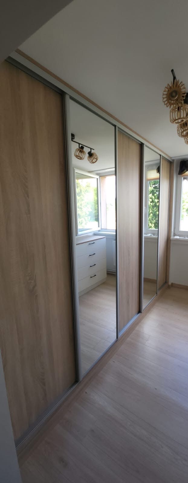 Jasna szafa wnękowa z przesuwnymi drzwiami z lustrami, odbijającymi wnętrze pokoju. Widoczna podłoga z paneli i komoda. Oświetlenie sufitowe w stylu boho.