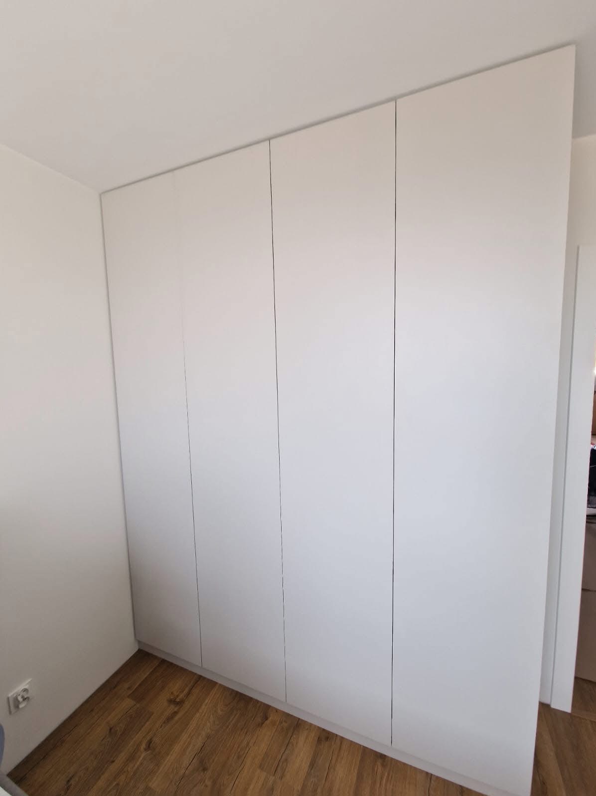 Minimalistyczna, biała szafa wnękowa z czterema drzwiami, idealnie wkomponowana w jasne wnętrze. Widoczna drewniana podłoga i fragment przejścia do innego pomieszczenia.