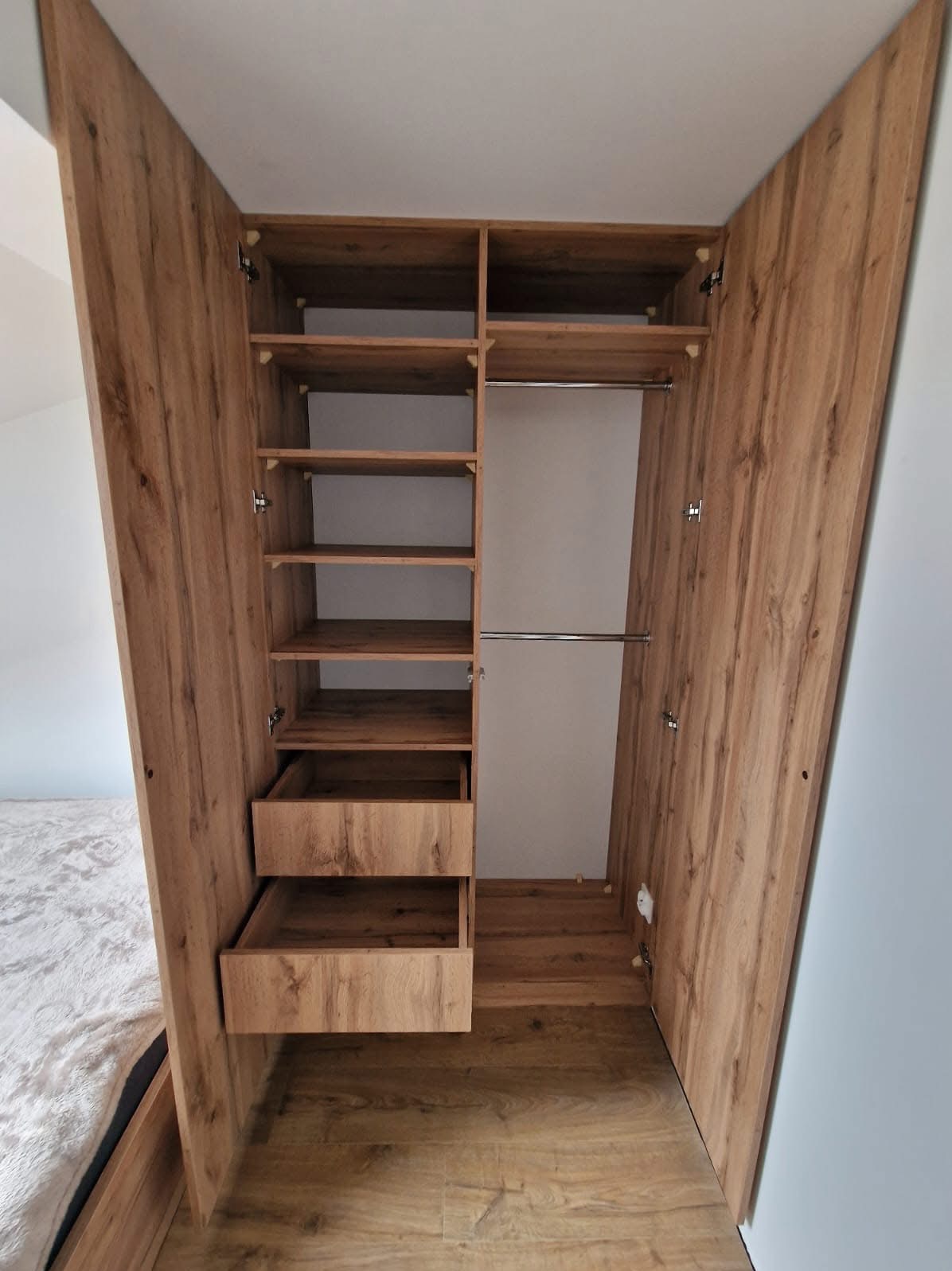 Otwarta szafa wnękowa z drewnianymi półkami i szufladami, widoczna z perspektywy podłogi, ukazująca wnętrze i strukturę drewna. Widoczne zawiasy i elementy montażowe.
