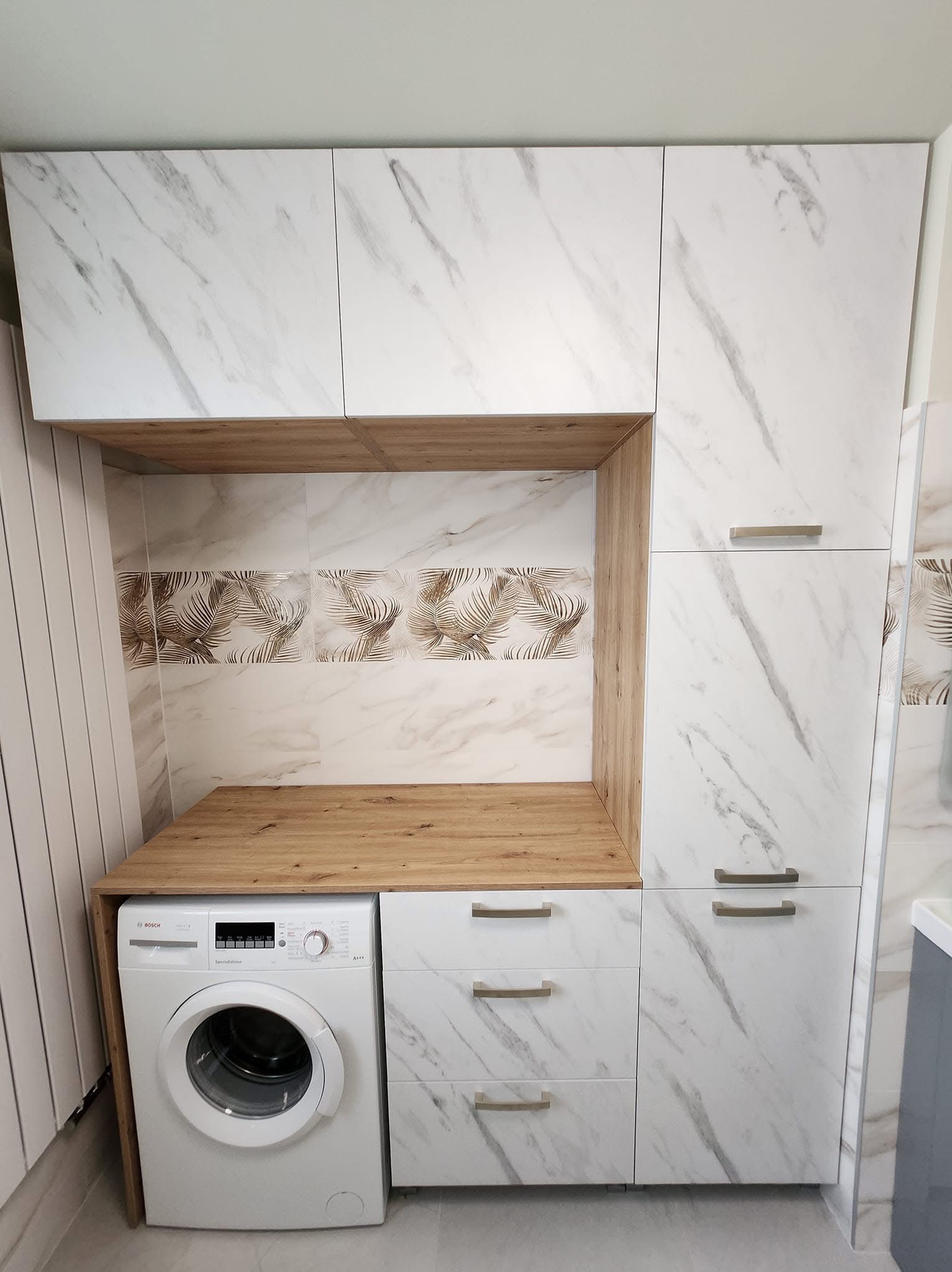 Biała zabudowa meblowa z marmurowym wzorem, integrująca pralkę. Blat drewniany, płytki z motywem liści palmowych. Nowoczesne uchwyty, minimalistyczny design.