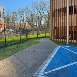 Szymon Łukasiewicz - Nowoczesne ogrodzenie panelowe w kolorze grafitowym, oddzielające parking od budynku mieszkalnego. Widoczna kostka brukowa i oznakowanie miejsca dla osób niepełnosprawnych.