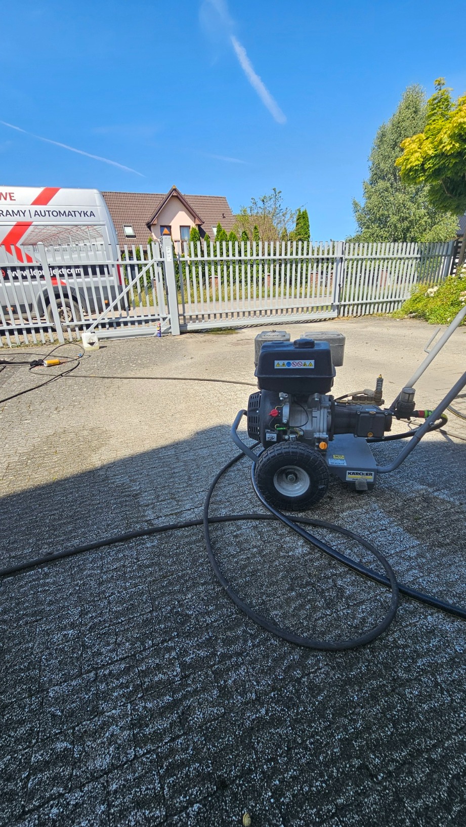 Myjka ciśnieniowa Karcher na kostce brukowej, w tle brama wjazdowa, budynek mieszkalny i firmowy bus z reklamą bram i automatyki. Słoneczny dzień.