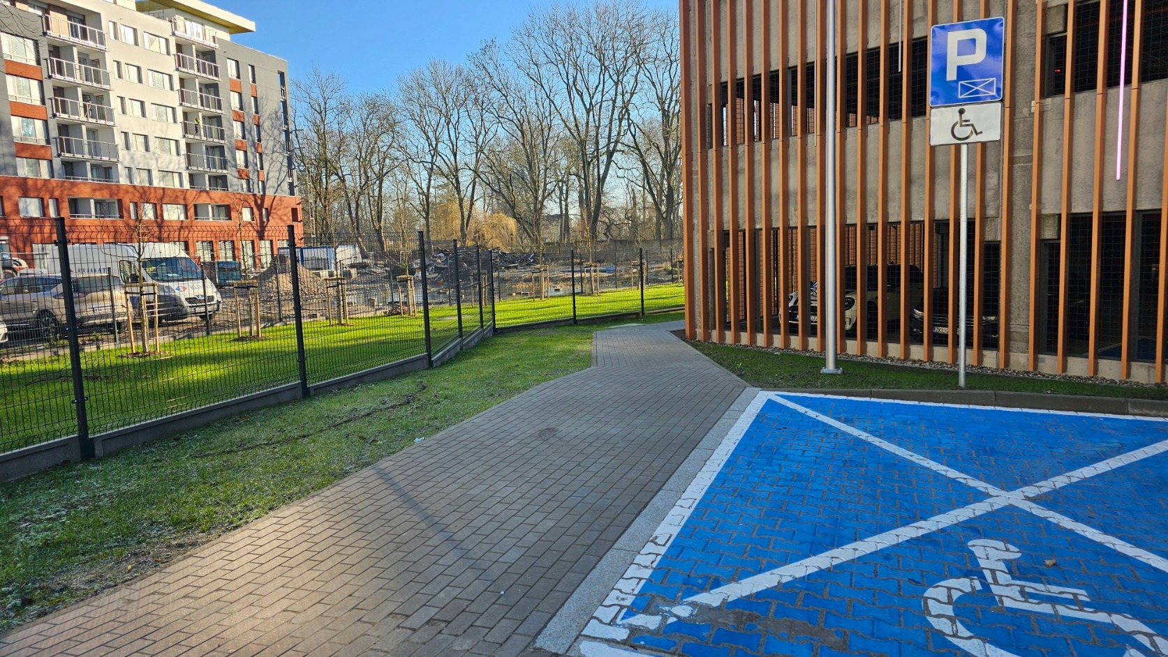 Nowoczesne ogrodzenie panelowe w kolorze grafitowym, oddzielające parking od budynku mieszkalnego. Widoczna kostka brukowa i oznakowanie miejsca dla osób niepełnosprawnych.