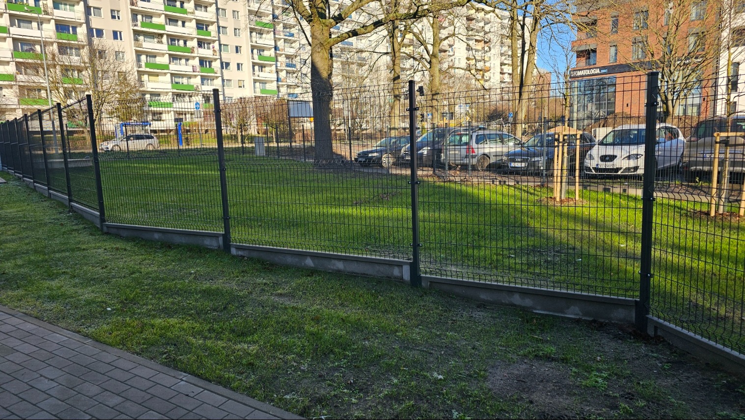 Nowoczesne ogrodzenie panelowe z podmurówką betonową na tle zielonego trawnika i zabudowy miejskiej. Widoczne słupki i siatka w kolorze grafitowym.