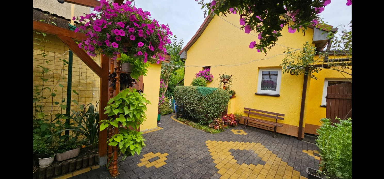 Żółty dom z kostką brukową po myciu, dekoracje kwiatowe, ławka. Kostka ułożona w geometryczny wzór, zadbana zieleń. Widok na budynek z perspektywy ogrodu.