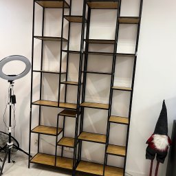 Witkowska Studio - Dwie wysokie, puste szafy regałowe z czarnymi metalowymi ramami i drewnianymi półkami, oświetlone lampą pierścieniową, stojące przy białej ścianie.