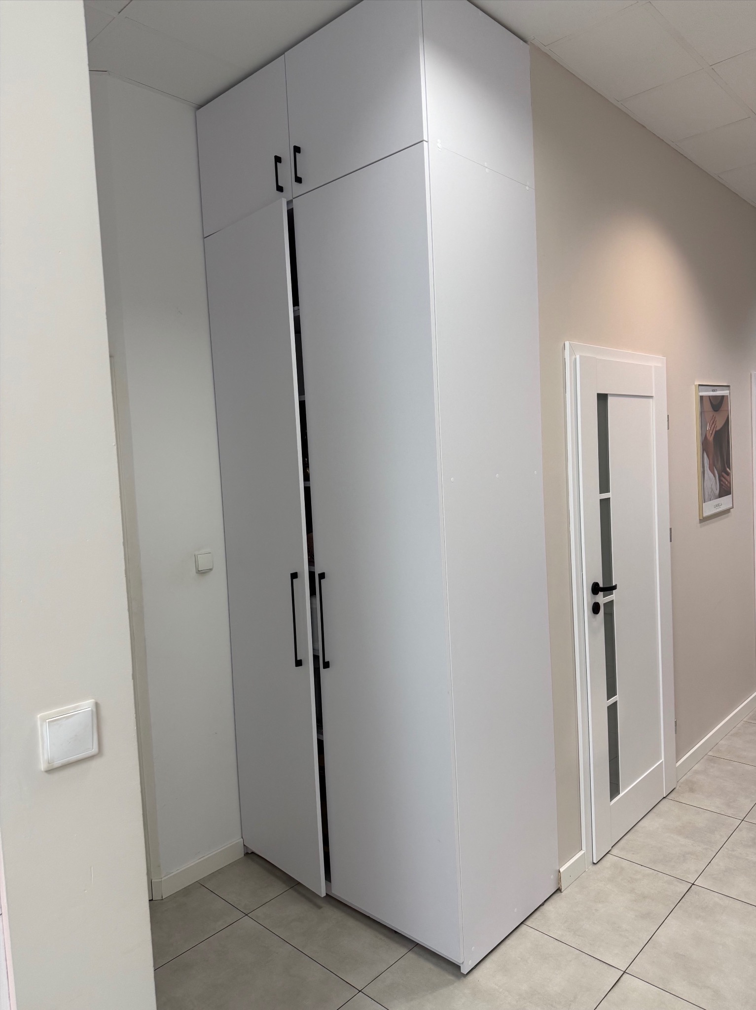 Wysoka, biała szafa z czarnymi uchwytami, częściowo otwarta, w jasnym wnętrzu. Widoczna podłoga z płytek i białe drzwi w tle. Minimalistyczny design.