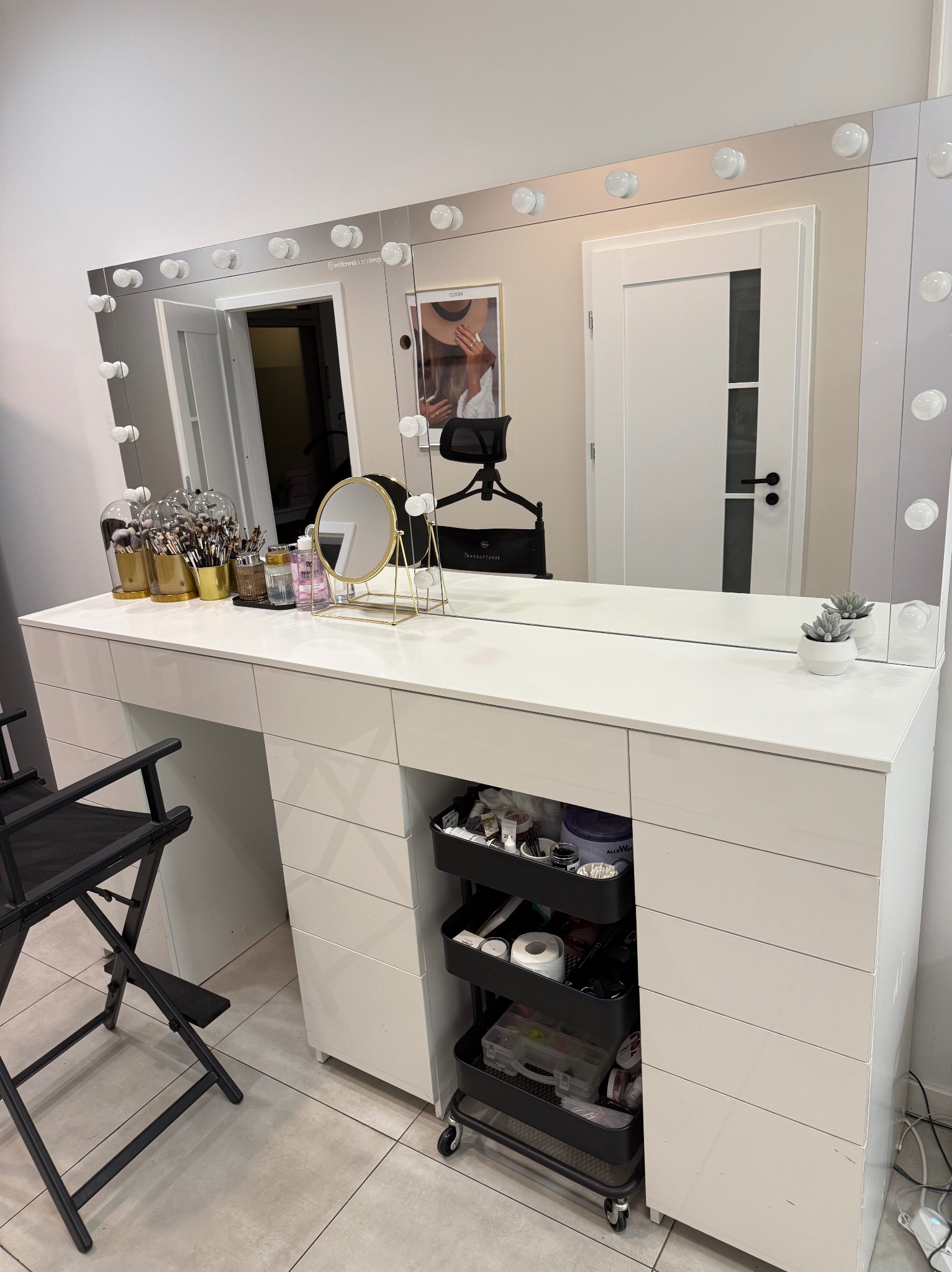 Biała toaletka z lustrem otoczonym żarówkami, czarne krzesło reżyserskie, kosmetyki i akcesoria do makijażu. Widoczne szuflady i wózek z kosmetykami.
