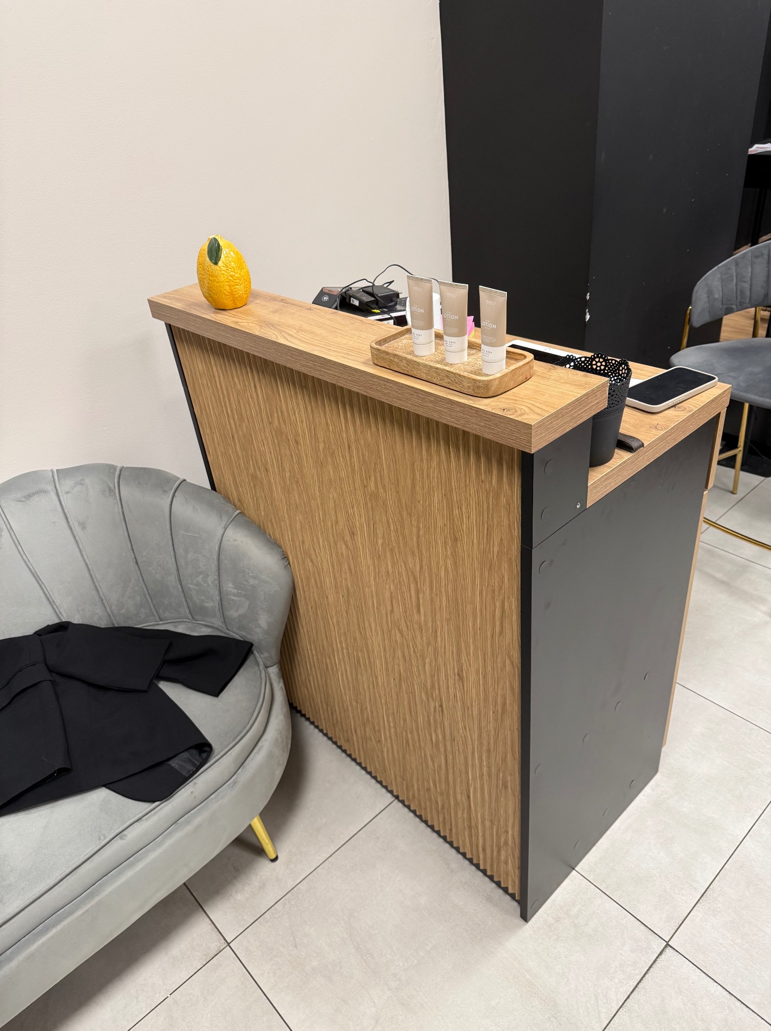 Recepcja z blatem imitującym drewno, szary fotel z czarną marynarką, dekoracje: ceramiczna cytryna i kosmetyki na tacy. Nowoczesny design, jasne wnętrze.