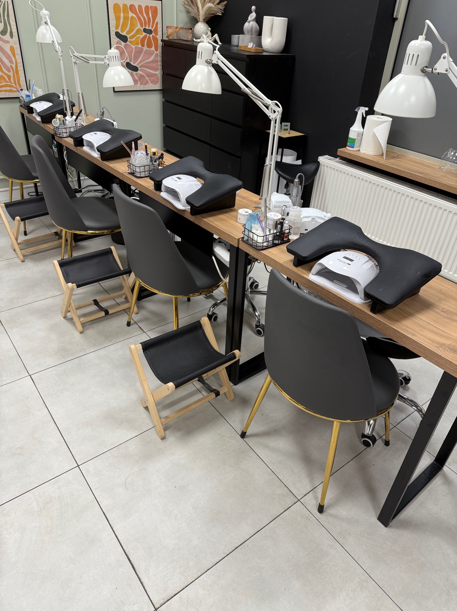 Nowoczesne stanowiska do manicure z lampami UV, czarnymi krzesłami na złotych nogach i drewnianymi blatami. Widok na salon kosmetyczny z dekoracjami na ścianie.