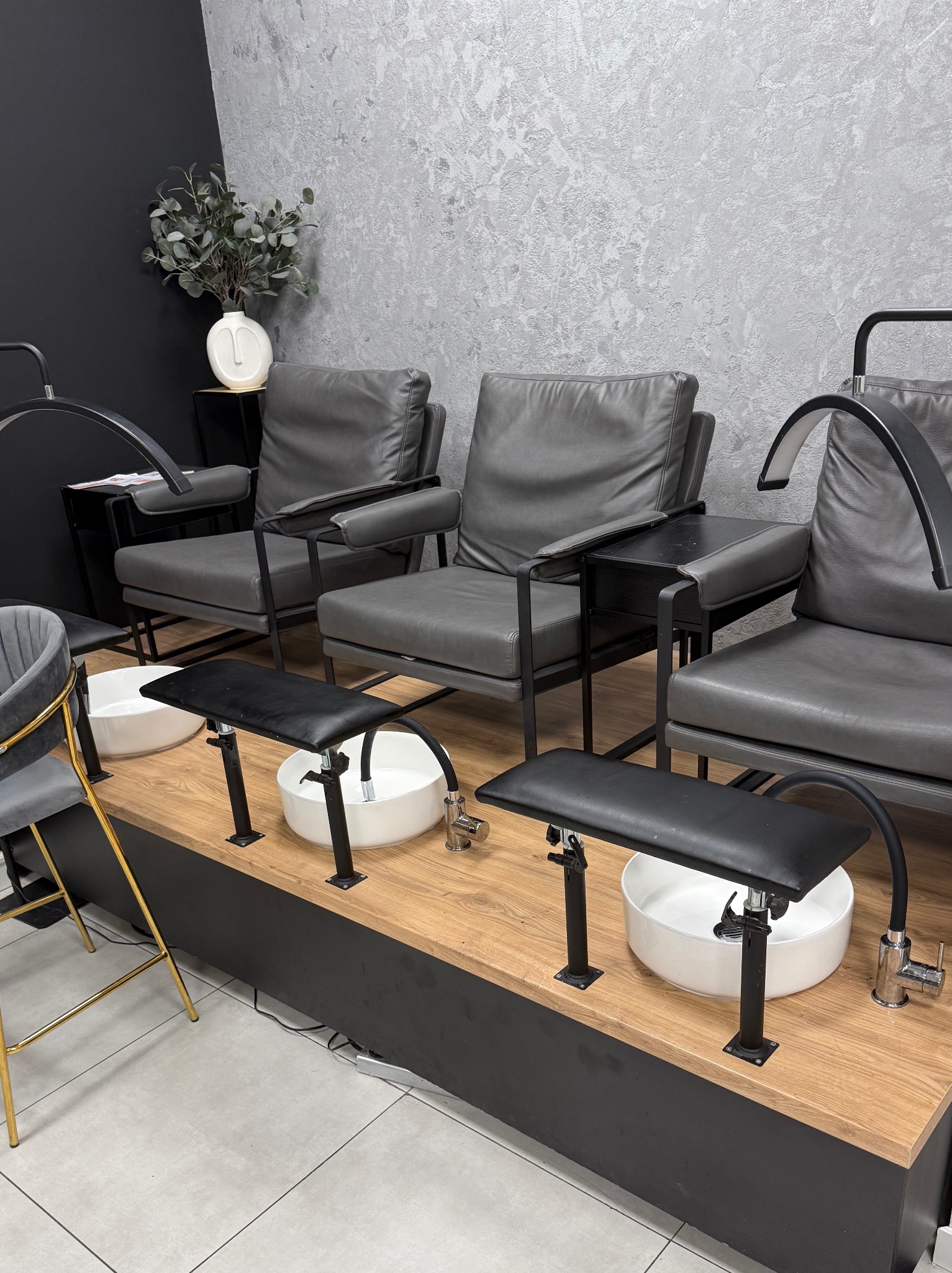 Nowoczesne stanowiska do pedicure z szarymi fotelami i białymi misami, oświetlone lampami. Minimalistyczny wystrój wnętrza w odcieniach szarości i czerni.
