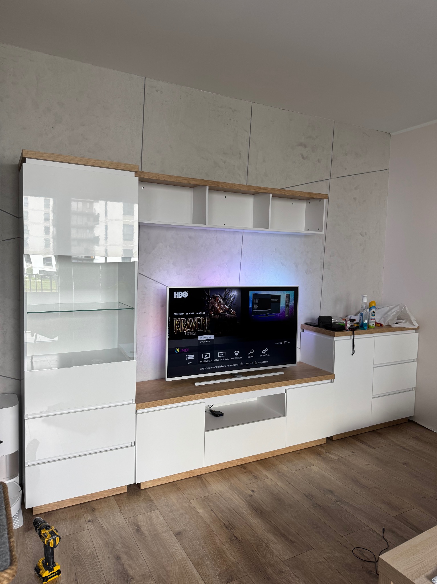 Biała meblościanka z drewnianymi akcentami, podświetlony telewizor, szara ściana z efektem betonu. W tle widoczne okno. Na podłodze leży wiertarka.