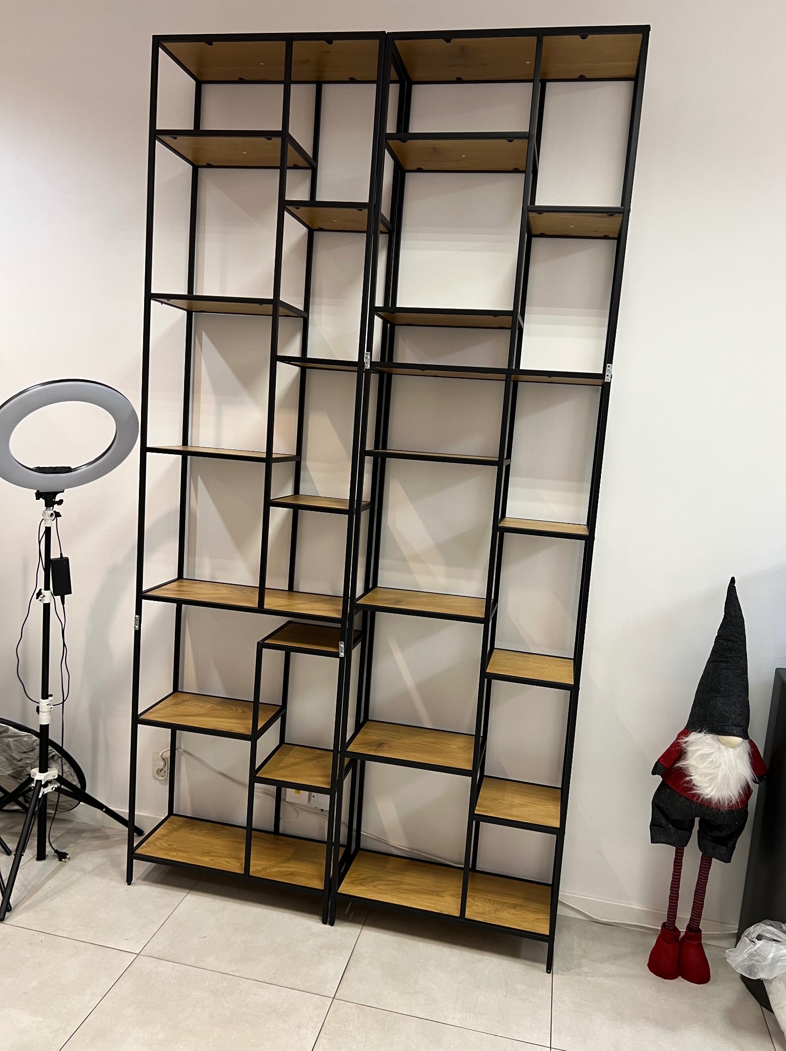Dwie wysokie, puste szafy regałowe z czarnymi metalowymi ramami i drewnianymi półkami, oświetlone lampą pierścieniową, stojące przy białej ścianie.