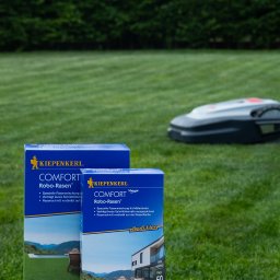 Fotografia Agata Wengerek - Dwa opakowania nasion trawy Kiepenkerl Comfort Robo-Rasen na tle trawnika i robota koszącego. Produkt na białym blacie, w tle zielony trawnik i żywopłot.