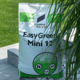 Fotografia Agata Wengerek - Worek nawozu Compo Expert EasyGreen Mini 12, ustawiony na betonowym murku, z zielonym trawnikiem w tle i ozdobnymi białymi kamieniami. Częściowo zasłonięty przez trawę.
