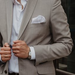 Fotografia Agata Wengerek - Mężczyzna w beżowym garniturze, biała koszula, złoty zegarek i obrączka. Widoczne dłonie zapinające marynarkę, biała poszetka w kieszonce. Elegancki, stonowany styl.