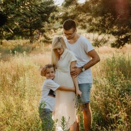 Fotografia Agata Wengerek - Rodzinne ujęcie w plenerze: przyszła mama w jasnej sukience, tata w okularach i syn tulą się, otoczeni wysoką trawą i drzewami w ciepłym, słonecznym świetle.
