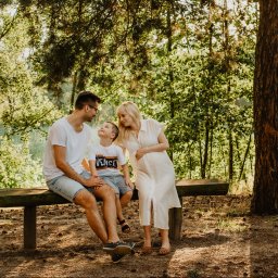 Fotografia Agata Wengerek - Rodzinne ujęcie w lesie: tata siedzi na ławce z synem, mama w białej sukience stoi obok, trzymając się za brzuszek. Ciepłe światło, naturalne tło.