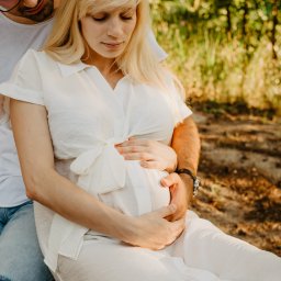 Fotografia Agata Wengerek - Jasnowłosa kobieta w białej sukience, siedząca na kolanach mężczyzny, obejmuje ciążowy brzuszek na tle zieleni. Delikatne światło podkreśla intymność i bliskość pary.