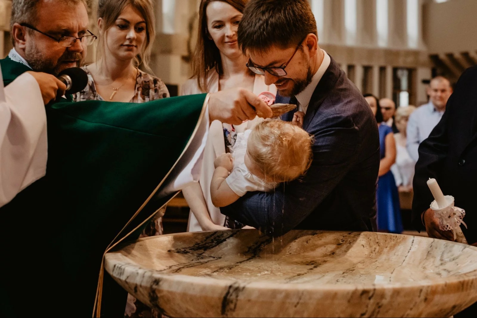 Chrzest dziecka w kościele: ksiądz polewa wodą główkę niemowlęcia trzymanego przez ojca. W tle rodzina i marmurowa chrzcielnica. Ujęcie z bliska, ciepłe światło.