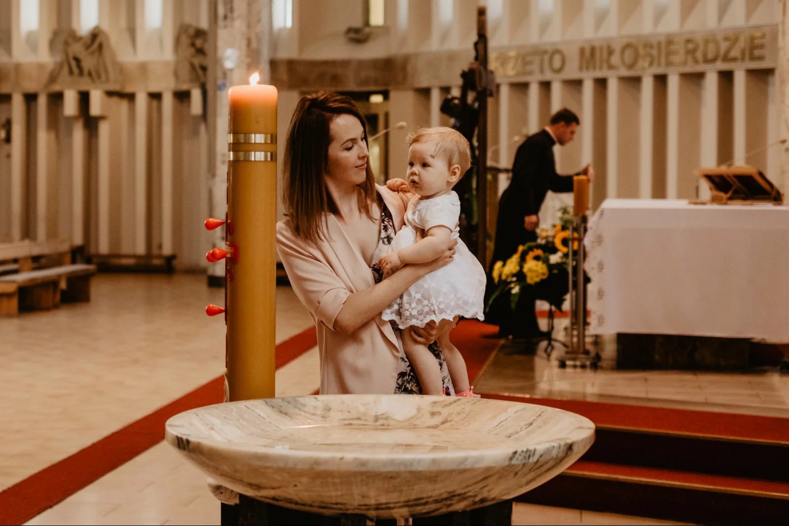 Matka trzymająca dziecko w białej sukience w kościele, obok chrzcielnica. W tle ołtarz i ksiądz. Ciepłe światło, uroczysta atmosfera.