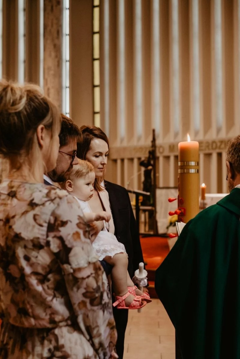Uroczystość Chrztu Świętego w kościele w Tarnowie Podgórnym. Rodzina z dzieckiem i ksiądz podczas ceremonii, w tle płonąca świeca i dekoracyjne ściany.