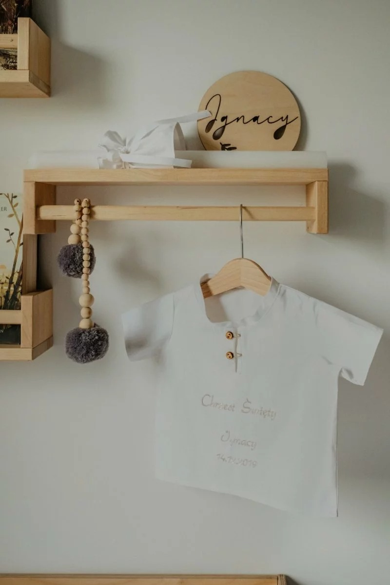 Biała koszulka z napisem 'Chrzest Święty Ignacy 14.11.2019' wisi na drewnianym wieszaku, obok drewniana ozdoba z imieniem 'Ignacy' i dekoracje w stylu boho.