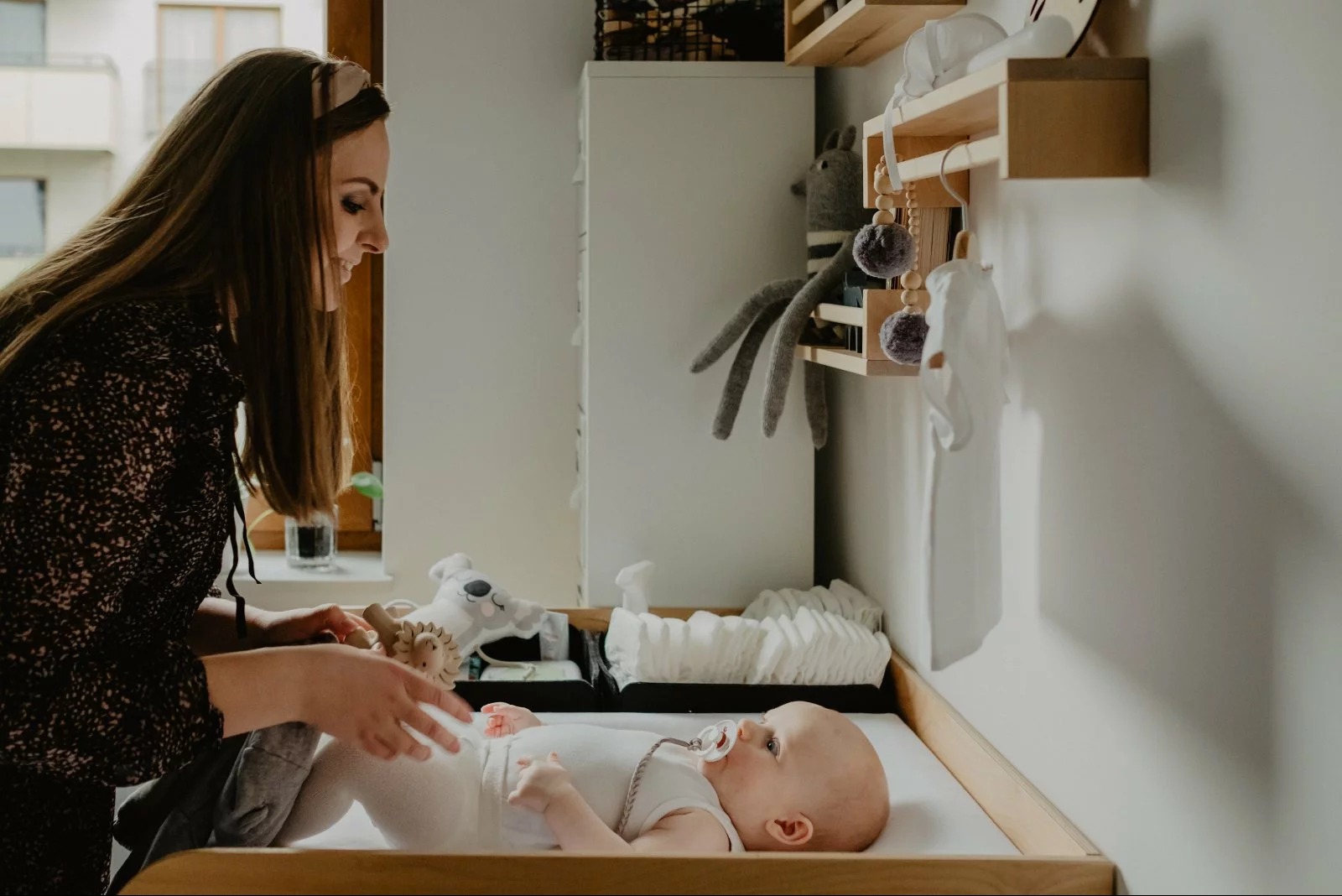 Mama pochyla się nad leżącym niemowlęciem na przewijaku w pokoju dziecięcym. Neutralne kolory, drewniane elementy, miękkie światło. Przygotowania do sesji.