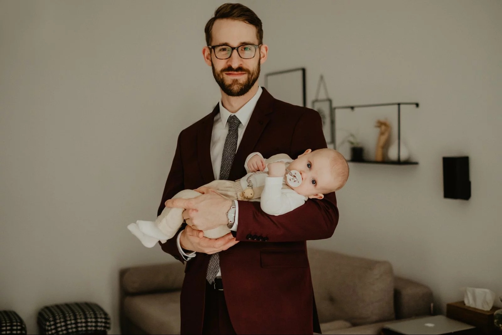 Elegancki mężczyzna w bordowym garniturze trzyma na rękach niemowlę z smoczkiem, w tle minimalistyczny salon. Sesja przed chrztem.