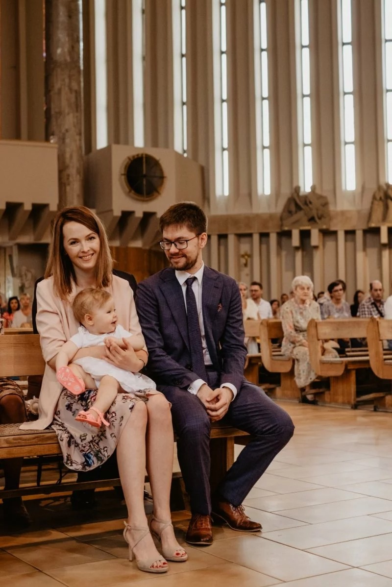 Uśmiechnięta rodzina siedzi na ławce w kościele. Matka trzyma dziecko, obok siedzi ojciec w garniturze. W tle widoczni inni uczestnicy uroczystości.