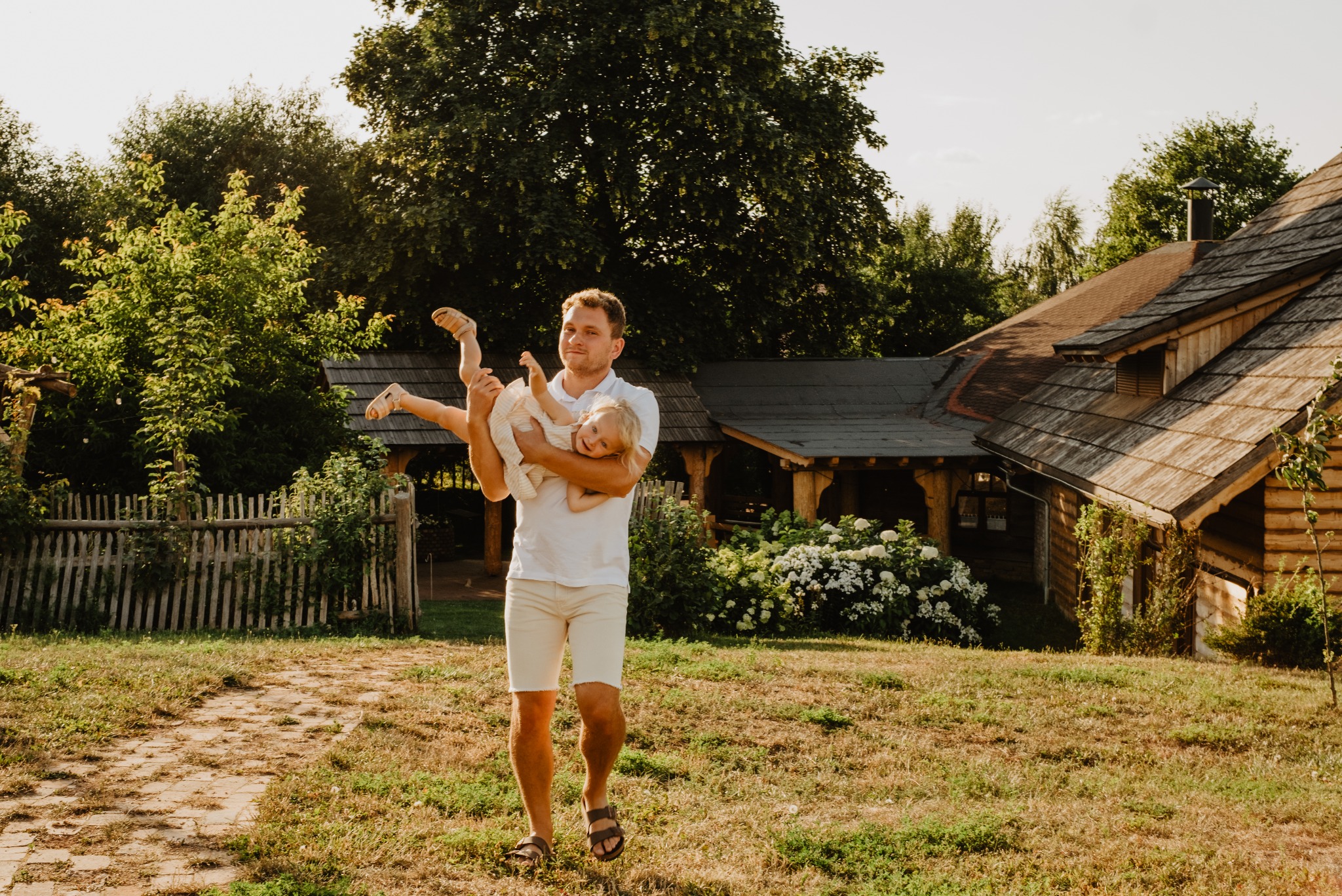 Uśmiechnięty tata w jasnych ubraniach podnosi córeczkę do góry na tle drewnianego domu i zieleni. Radosna, rodzinna atmosfera w letnim słońcu.
