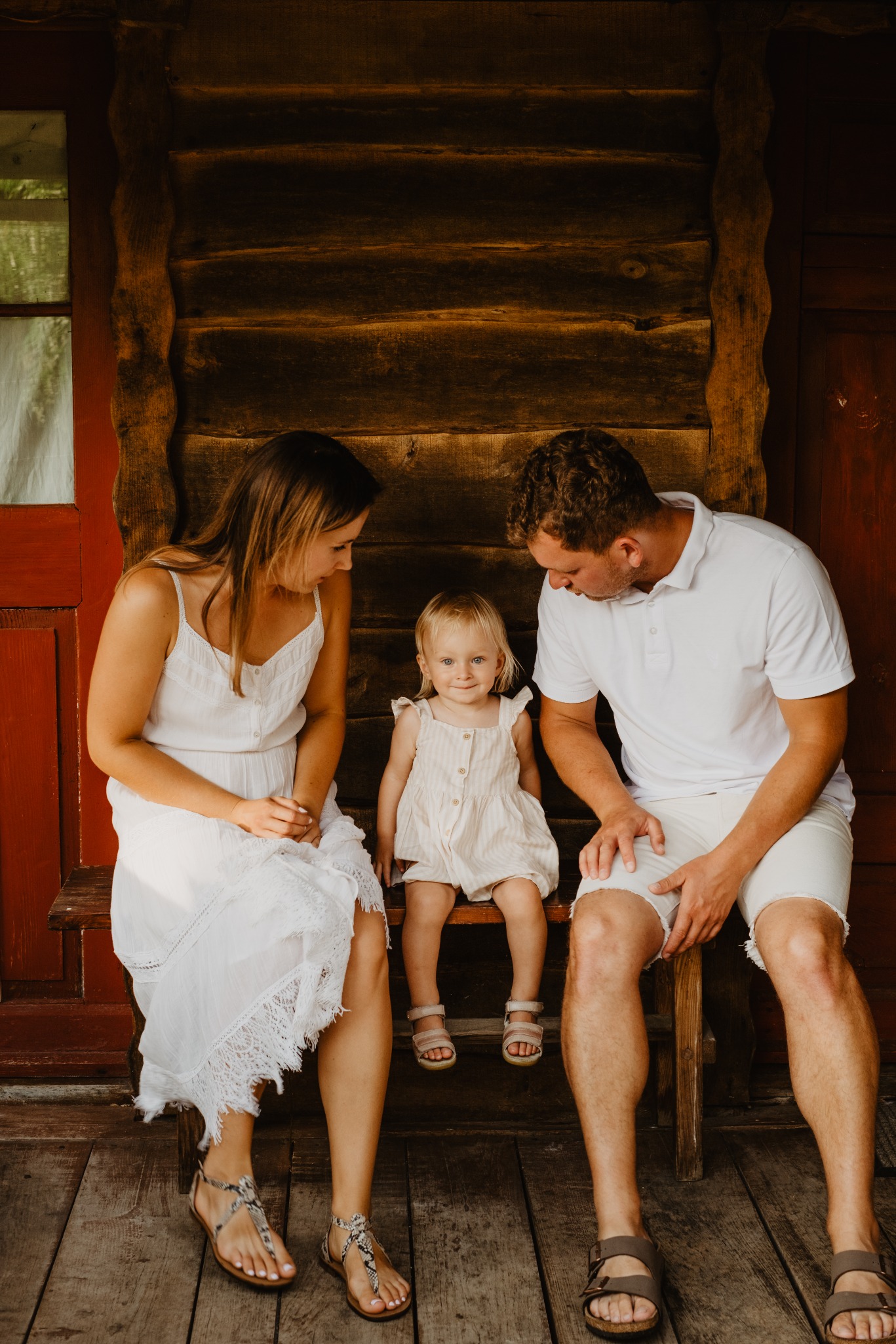 Rodzinna fotografia: Mama, tata i córka siedzą na drewnianej ławce na tle rustykalnej ściany z bali. Ubrani w jasne stroje, patrzą na siebie z czułością.